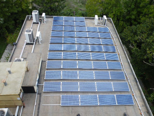 Widok z góry na instalację paneli słonecznych na płaskim dachu budynku, z widocznymi jednostkami klimatyzacji na obrzeżach dachu i zielenią drzew w tle.