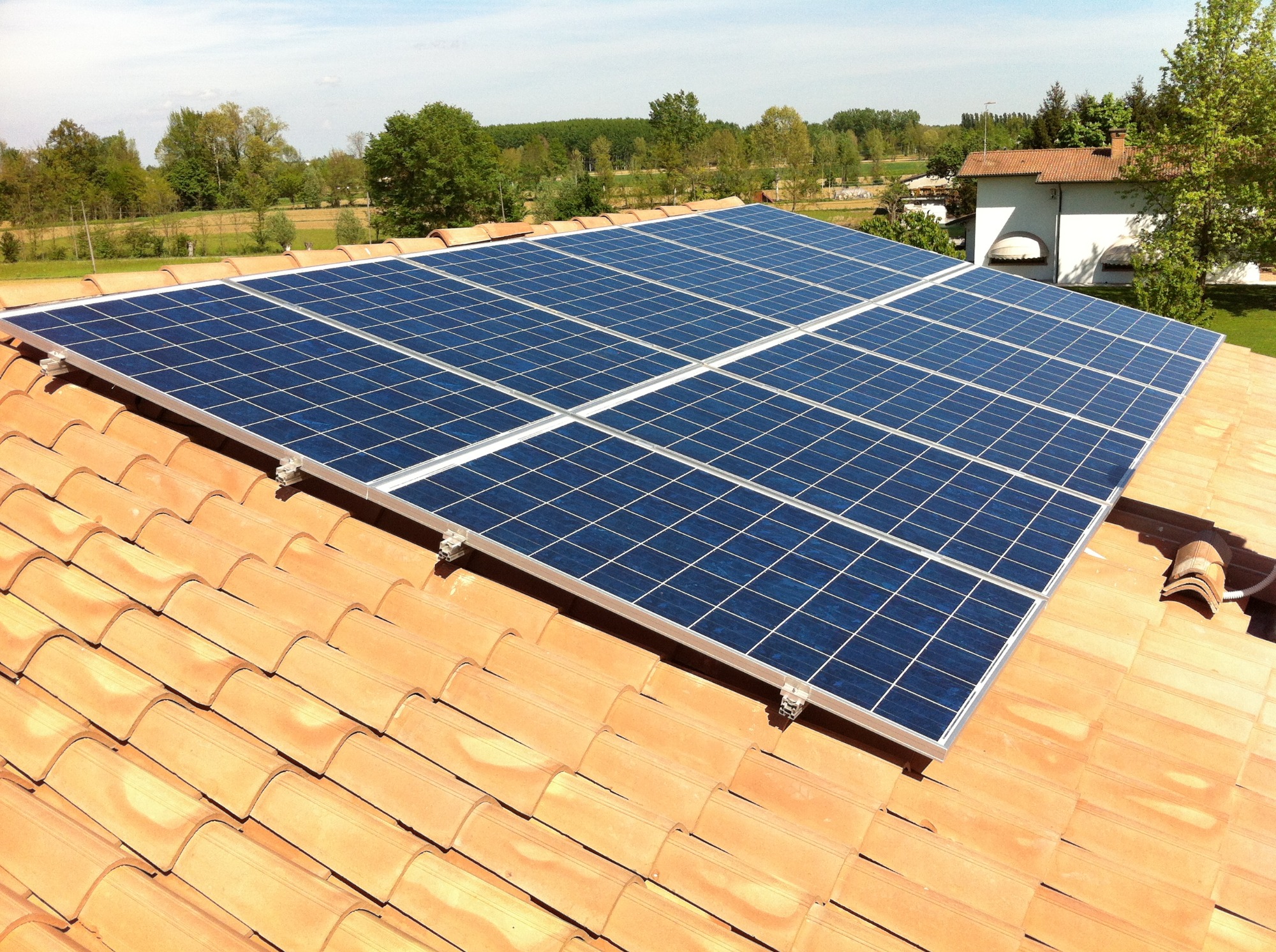 Panele słoneczne zamontowane na dachu z pomarańczowej dachówki, widok z góry, w tle budynek i drzewa.