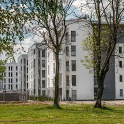 Nowoczesny, wielopiętrowy budynek mieszkalny z białymi elewacjami i dużymi oknami, widoczny z perspektywy trawnika z drzewami na pierwszym planie, przy słonecznej pogodzie.