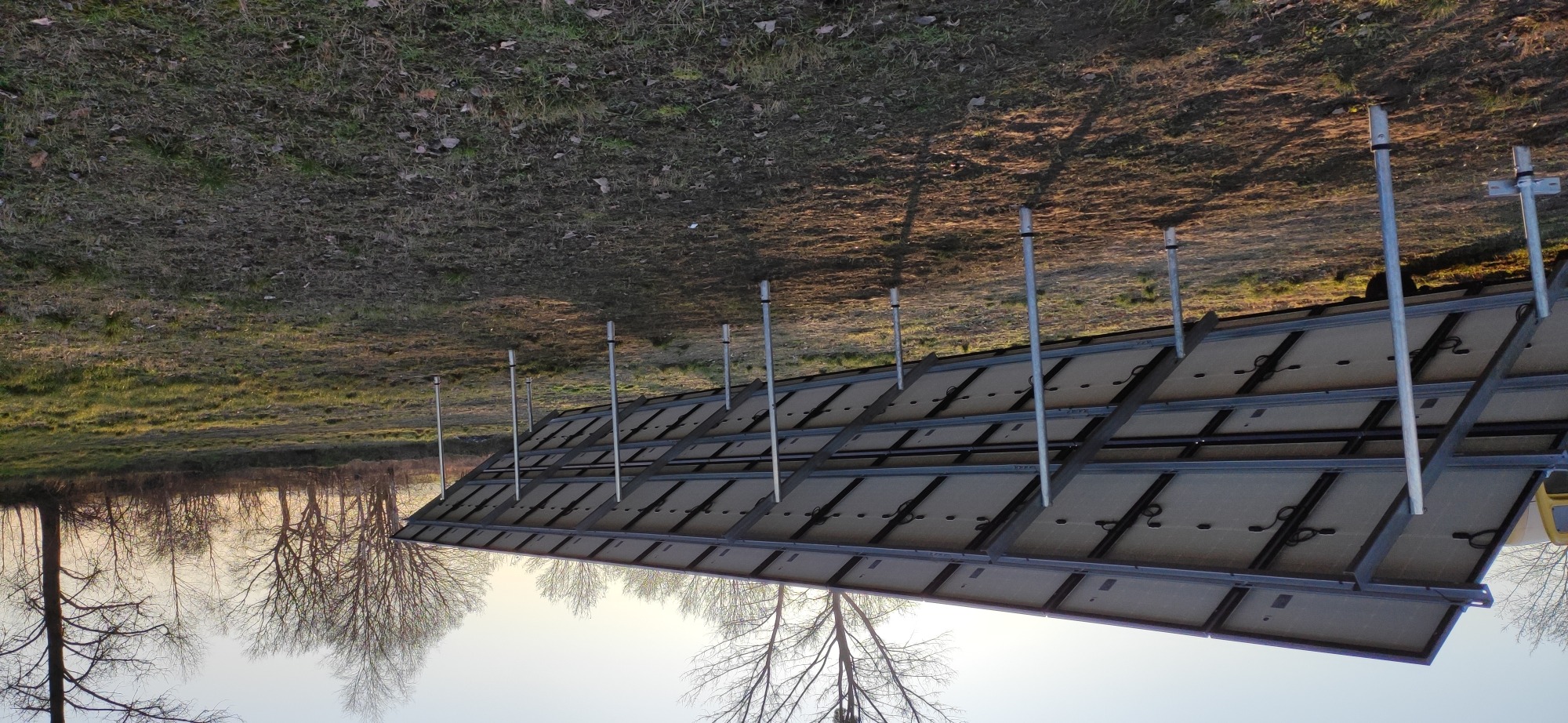 Widok od spodu na panele słoneczne zamontowane na metalowej konstrukcji z rurami, na tle trawy i drzew bez liści.