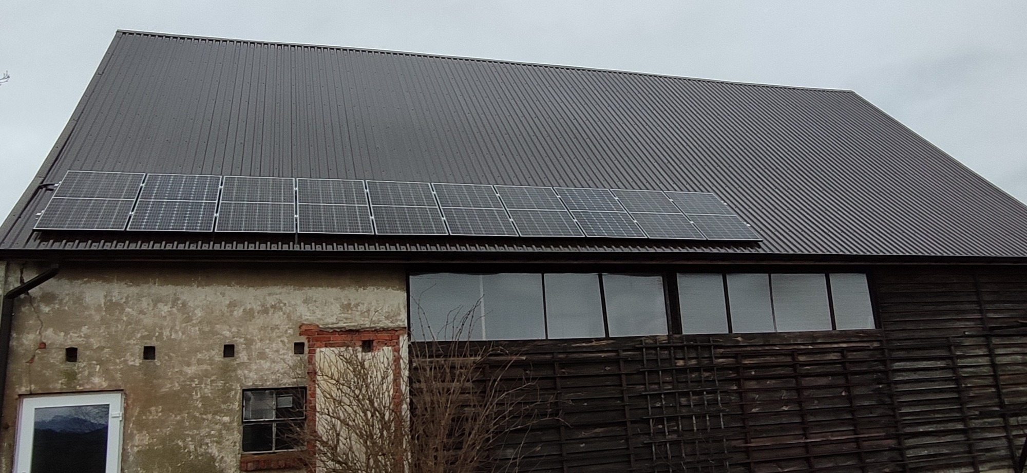 Panele słoneczne zamontowane na dachu starego, częściowo drewnianego budynku gospodarczego z widocznymi śladami użytkowania.