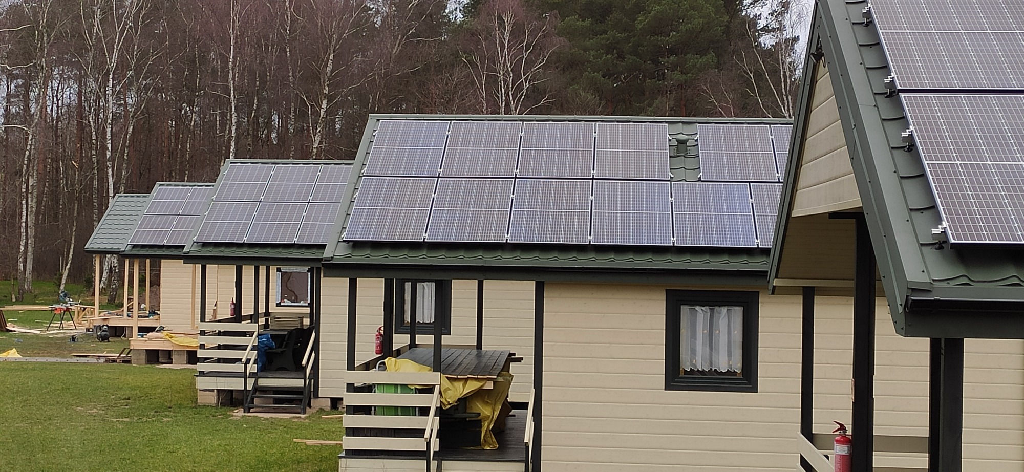 Rząd domków letniskowych z panelami fotowoltaicznymi na dachach, widoczne narzędzia budowlane przy jednym z domków, las w tle.
