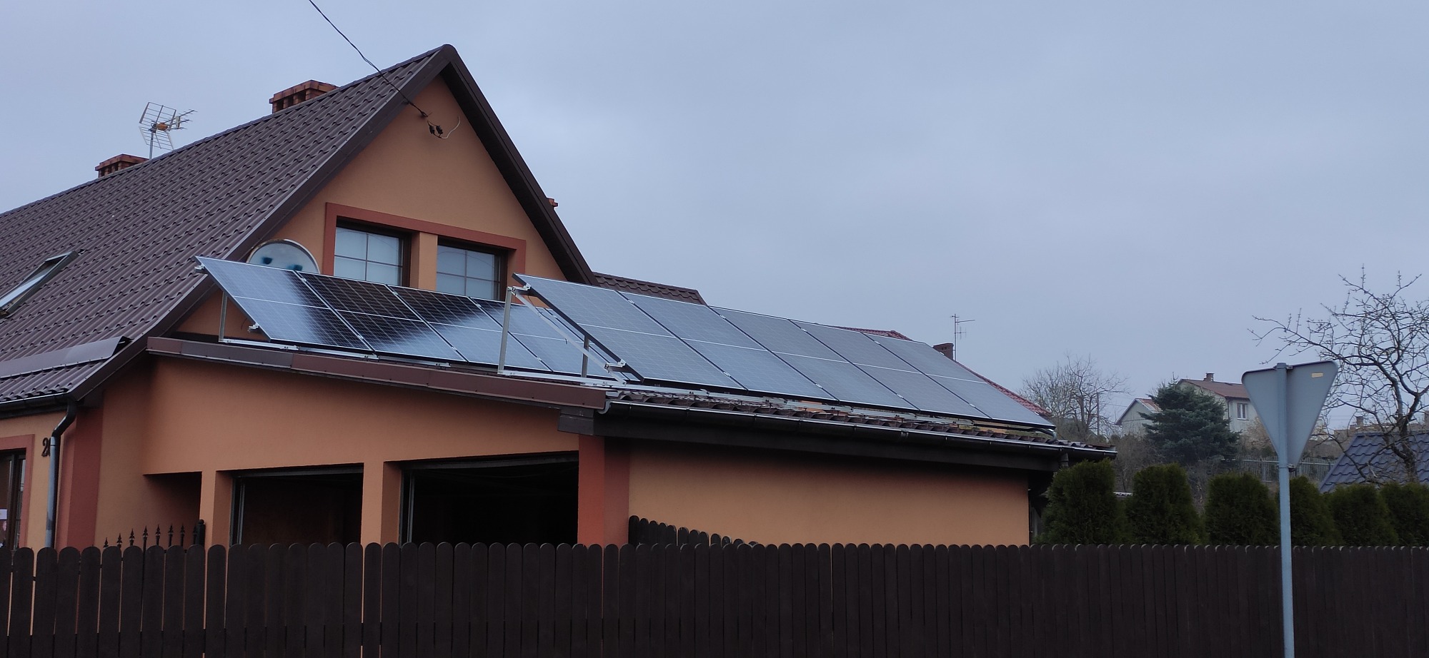 Panele słoneczne zamontowane na dachu domu jednorodzinnego z brązową dachówką, widoczne zza drewnianego ogrodzenia. Część paneli umieszczona na wspornikach.