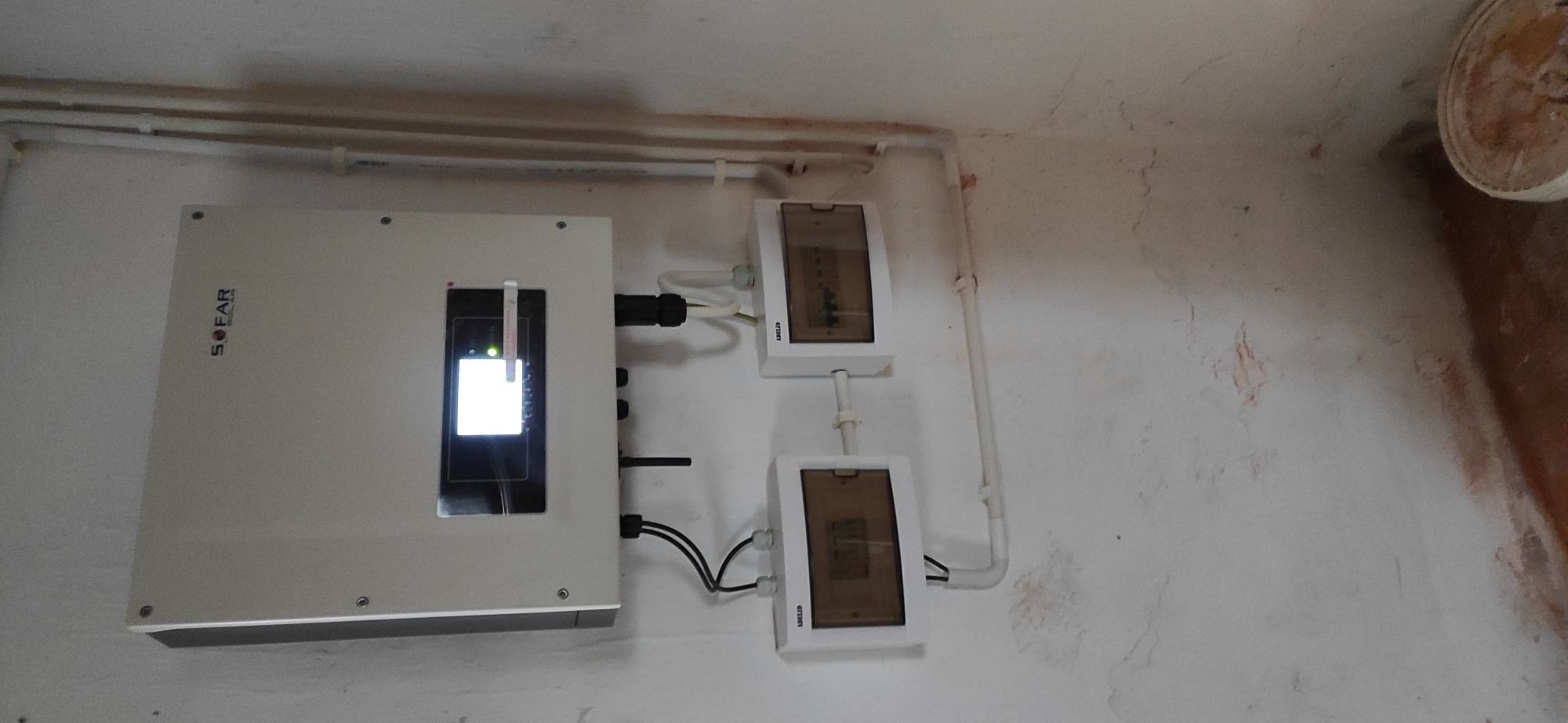 Zamontowany inwerter Sofar Solar z podłączonymi przewodami i dwiema skrzynkami zabezpieczeń na ścianie wewnątrz budynku.