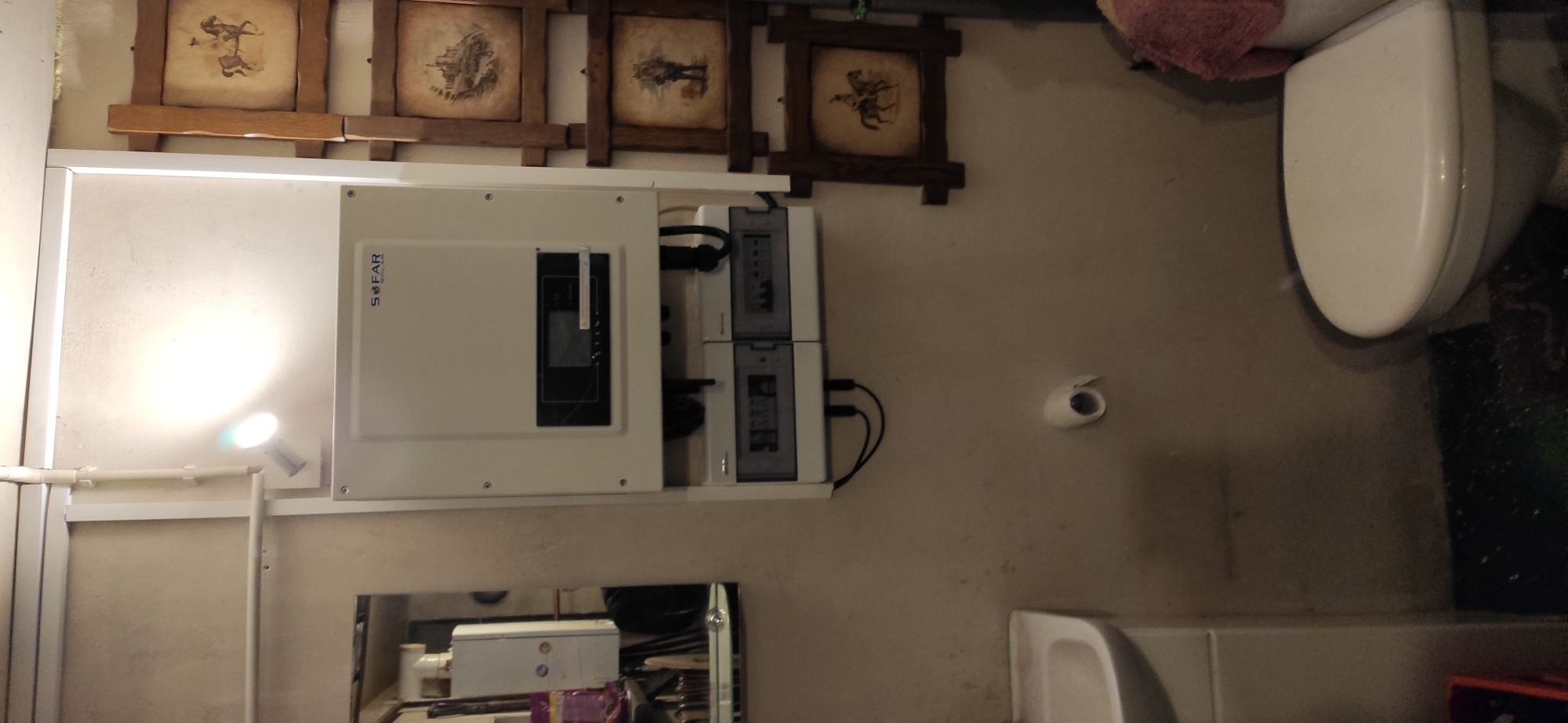 Biała skrzynka z logo SOFAR i aparaturą elektryczną zamontowana na ścianie w pomieszczeniu, widoczne elementy instalacji elektrycznej i fragment toalety.