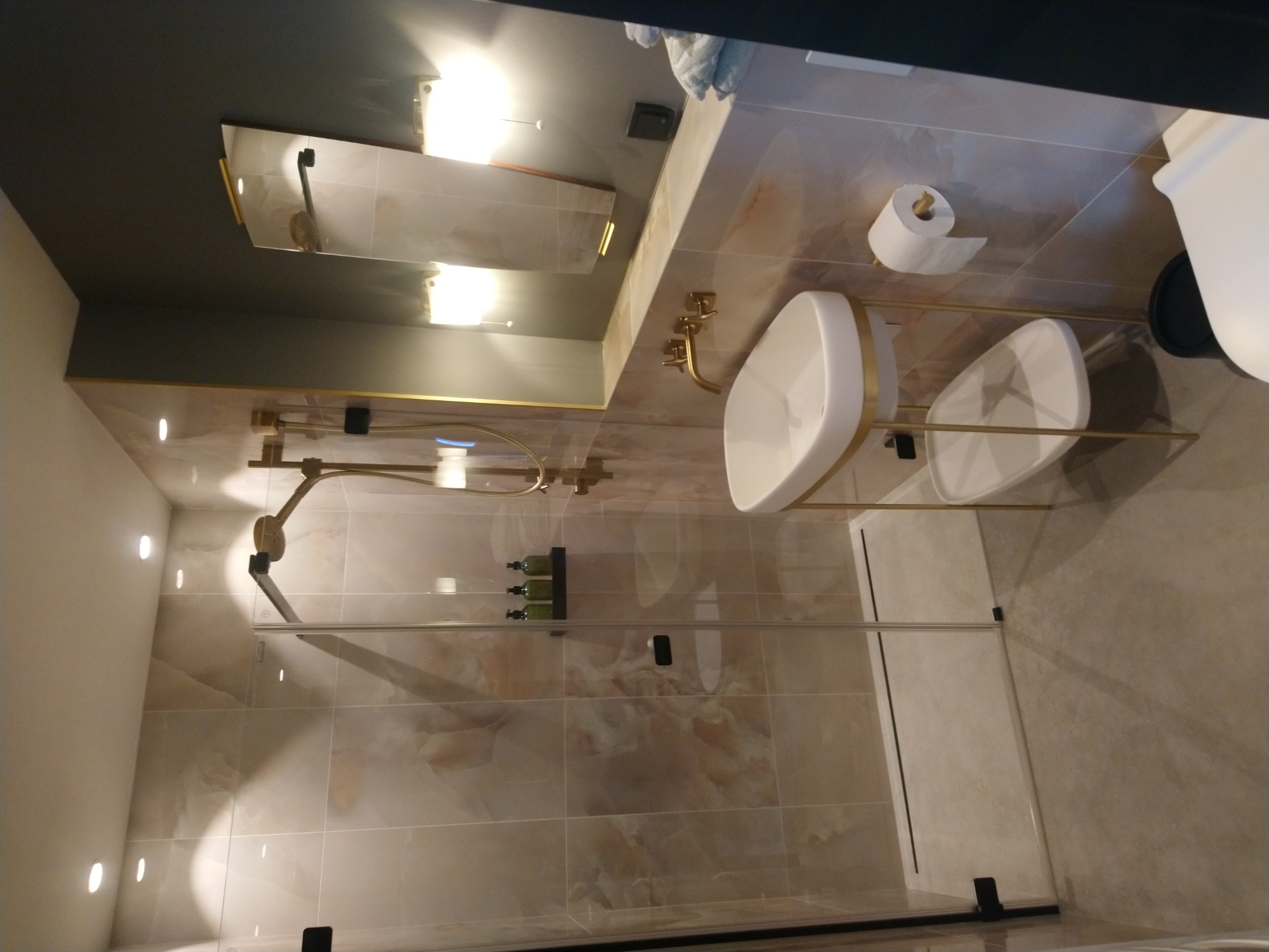 Nowoczesna łazienka z kabiną prysznicową typu walk-in, umywalką nablatową na złotym stelażu, armaturą w kolorze złota i marmurowymi płytkami na ścianach.