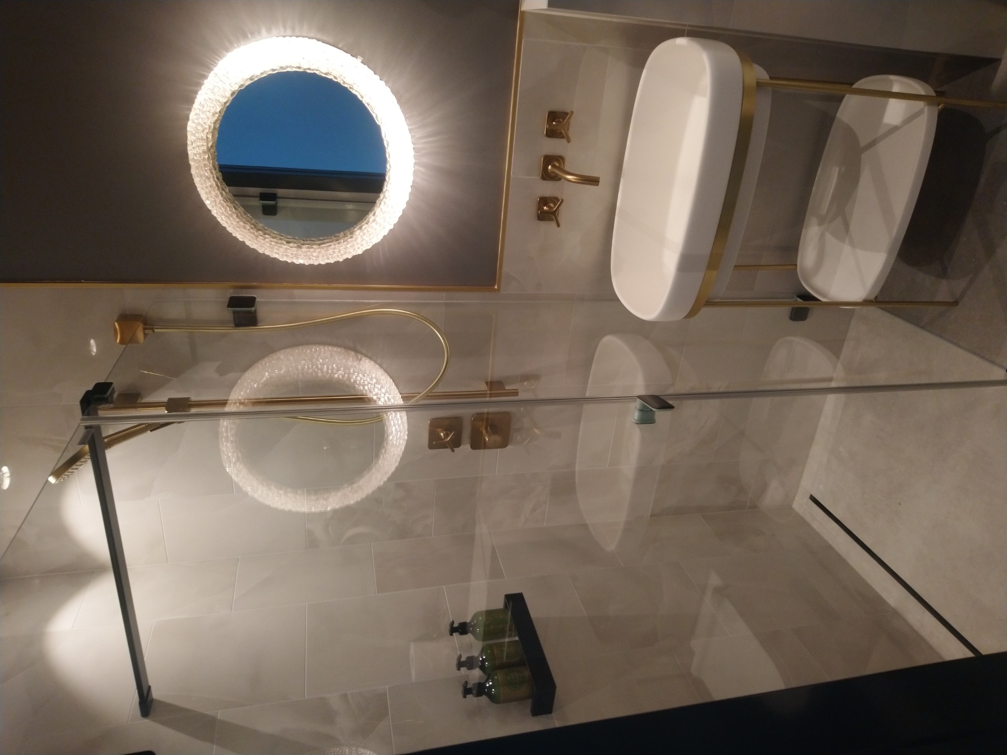 Nowoczesna łazienka z okrągłym lustrem z podświetleniem LED, szklaną kabiną prysznicową, złotą armaturą i półkami na kosmetyki w odcieniach bieli i szarości.