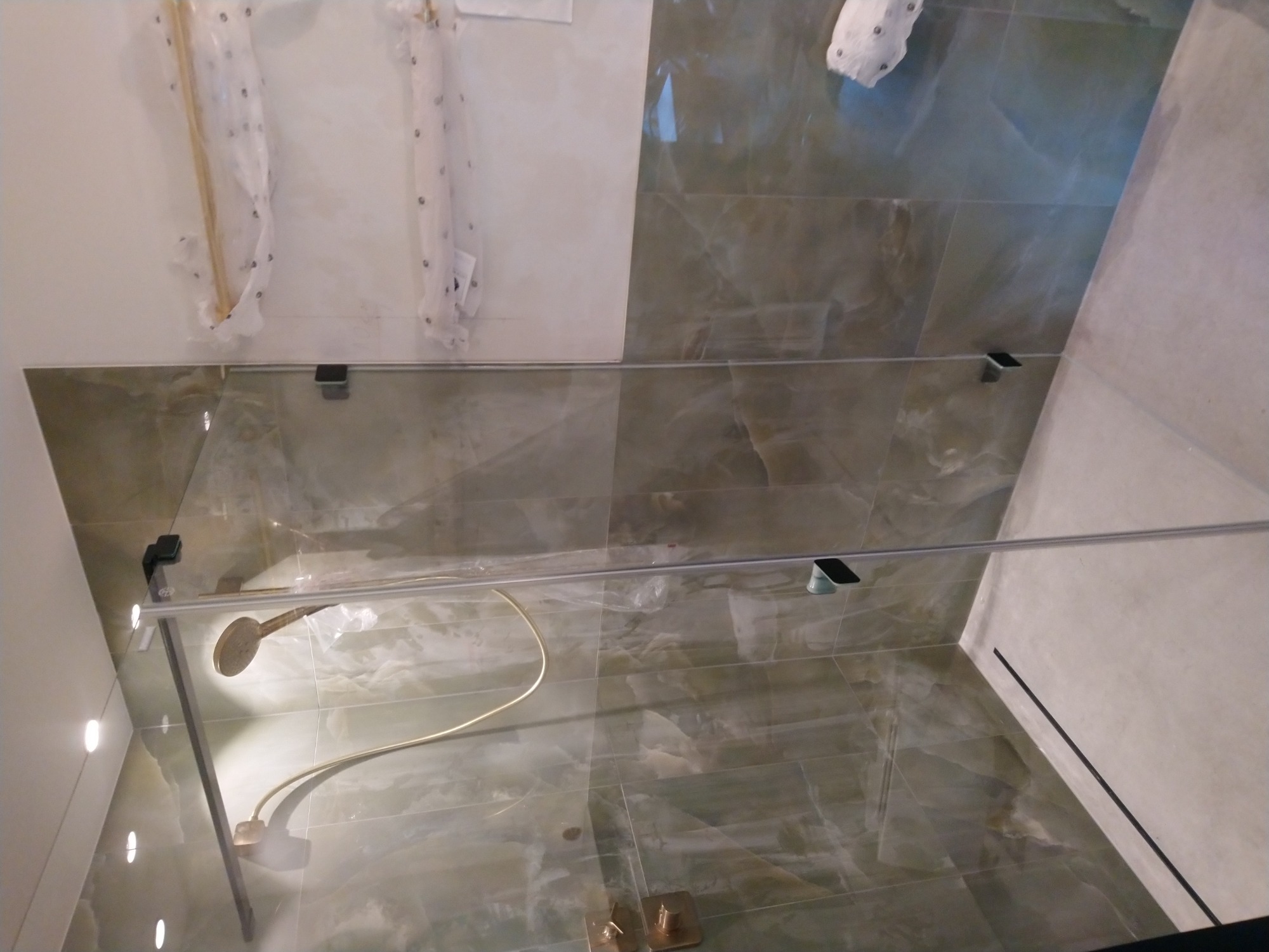 Szklana kabina prysznicowa z zamontowaną deszczownicą na tle zielonych płytek imitujących marmur, widoczne elementy montażowe i zabezpieczające folie na ścianach.