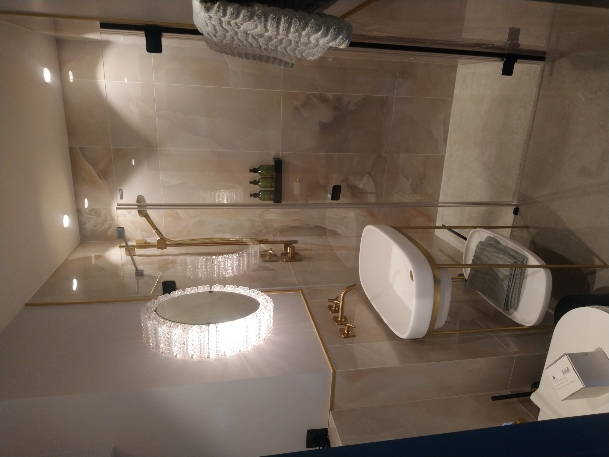 Nowoczesna łazienka z beżowymi płytkami imitującymi marmur, kabina prysznicowa ze złotymi elementami, umywalka nablatowa z baterią w kolorze złota, okrągłe lustro z kryształkami i szary ręcznik.