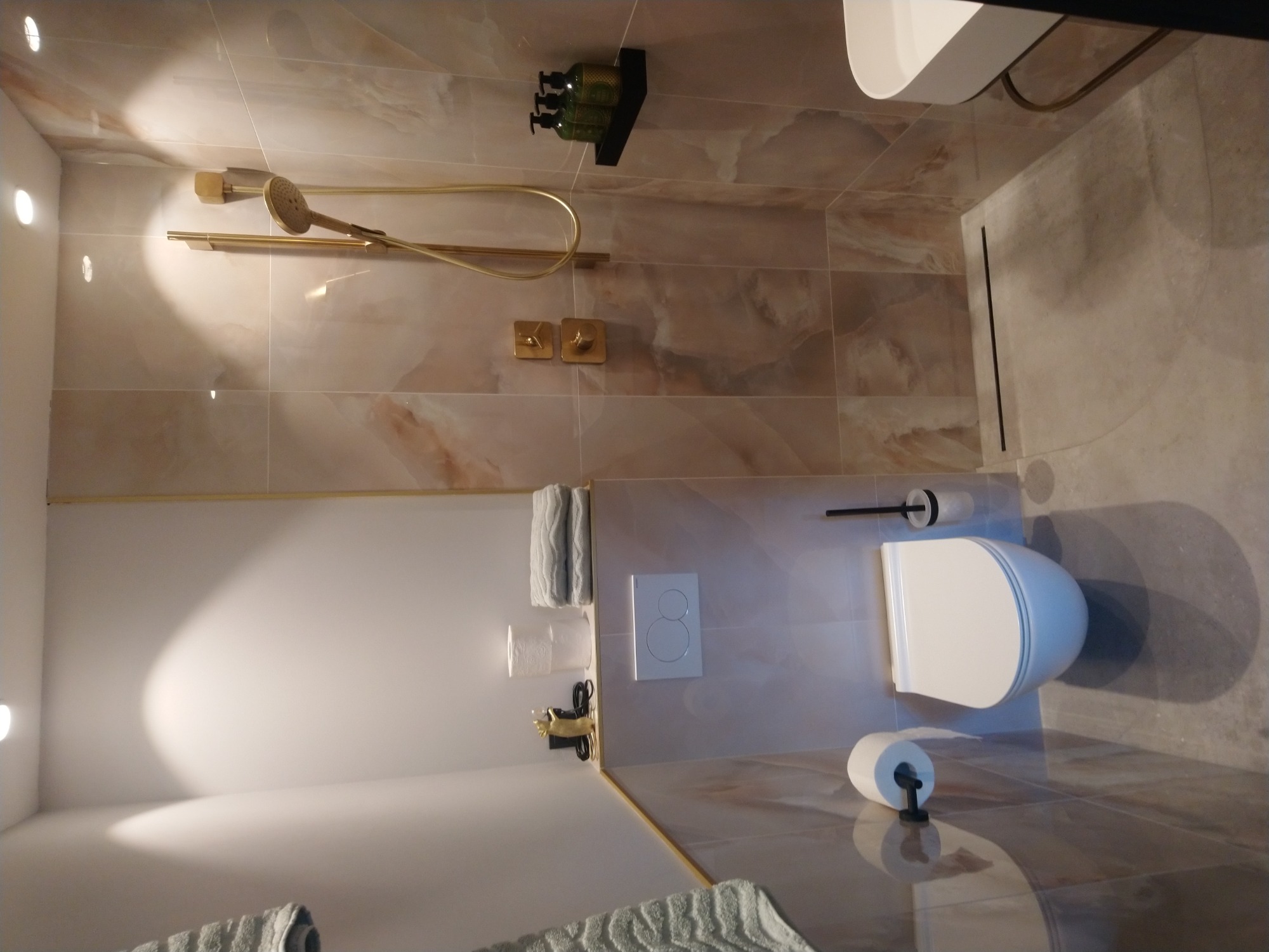 Nowoczesna łazienka z beżowymi płytkami imitującymi marmur, złotą armaturą prysznicową, podwieszaną toaletą i akcesoriami w kolorze czarnym i złotym.