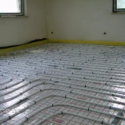 Ułożone rury ogrzewania podłogowego na folii z czerwoną kratką, widoczne okna i fragment ściany. Na ścianie widoczne gniazdka elektryczne.