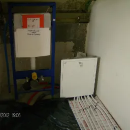 Stelaż WC JOMO w niebieskiej ramie, biała obudowa z instrukcją, rury ogrzewania podłogowego, biała szafka z kluczem, surowy stan wykończenia ścian.