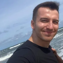 Uśmiechnięty mężczyzna w czarnej koszulce na tle morza i pochmurnego nieba, z widocznymi śladami po bańkach na szyi.