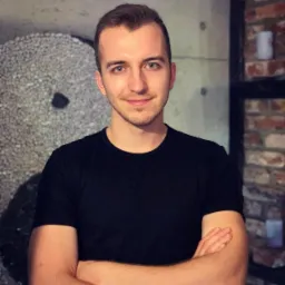 Portret młodego mężczyzny z niebieskimi oczami, uśmiechniętego, w czarnej koszulce, z rękami skrzyżowanymi na piersiach, na tle dekoracyjnej ściany z kamienia i cegły.