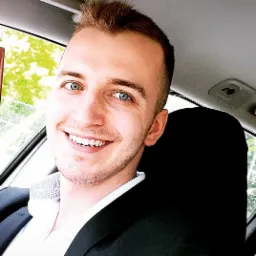 Portret uśmiechniętego mężczyzny w garniturze, siedzącego w samochodzie, z widocznym fragmentem zieleni za oknem.