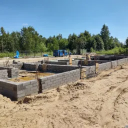 Wykopane fundamenty pod budynek z częściowo wymurowanymi ścianami z betonowych bloczków, wystającymi prętami zbrojeniowymi, na piaszczystym terenie budowy z widocznym niebieskim busem w tle.