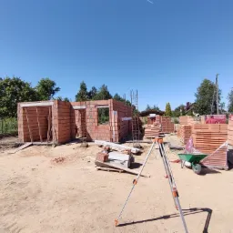 Wznoszenie ścian parterowego budynku z czerwonej cegły ceramicznej, widoczne zbrojenia pionowe, zapas materiału budowlanego na paletach oraz taczka, w tle zieleń drzew i błękitne niebo.