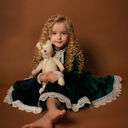 Małgorzata Popęda Fotografie - Dziewczynka z kręconymi blond włosami, ubrana w zieloną, aksamitną sukienkę z koronkowym wykończeniem, siedzi na brązowym tle, trzymając pluszowego misia. Jedna stopa jest widoczna na pierwszym...