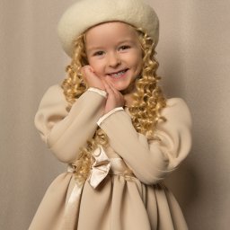 Małgorzata Popęda Fotografie - Portret uśmiechniętej dziewczynki z blond lokami w beżowej sukience z kokardą i wełnianym berecie na tle beżowej ściany.