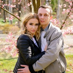 Małgorzata Popęda Fotografie - Para obejmująca się pod kwitnącym drzewem wiśni, kobieta w czarnej kurtce i jeansach, mężczyzna w beżowej kurtce, delikatne różowe kwiaty w tle.