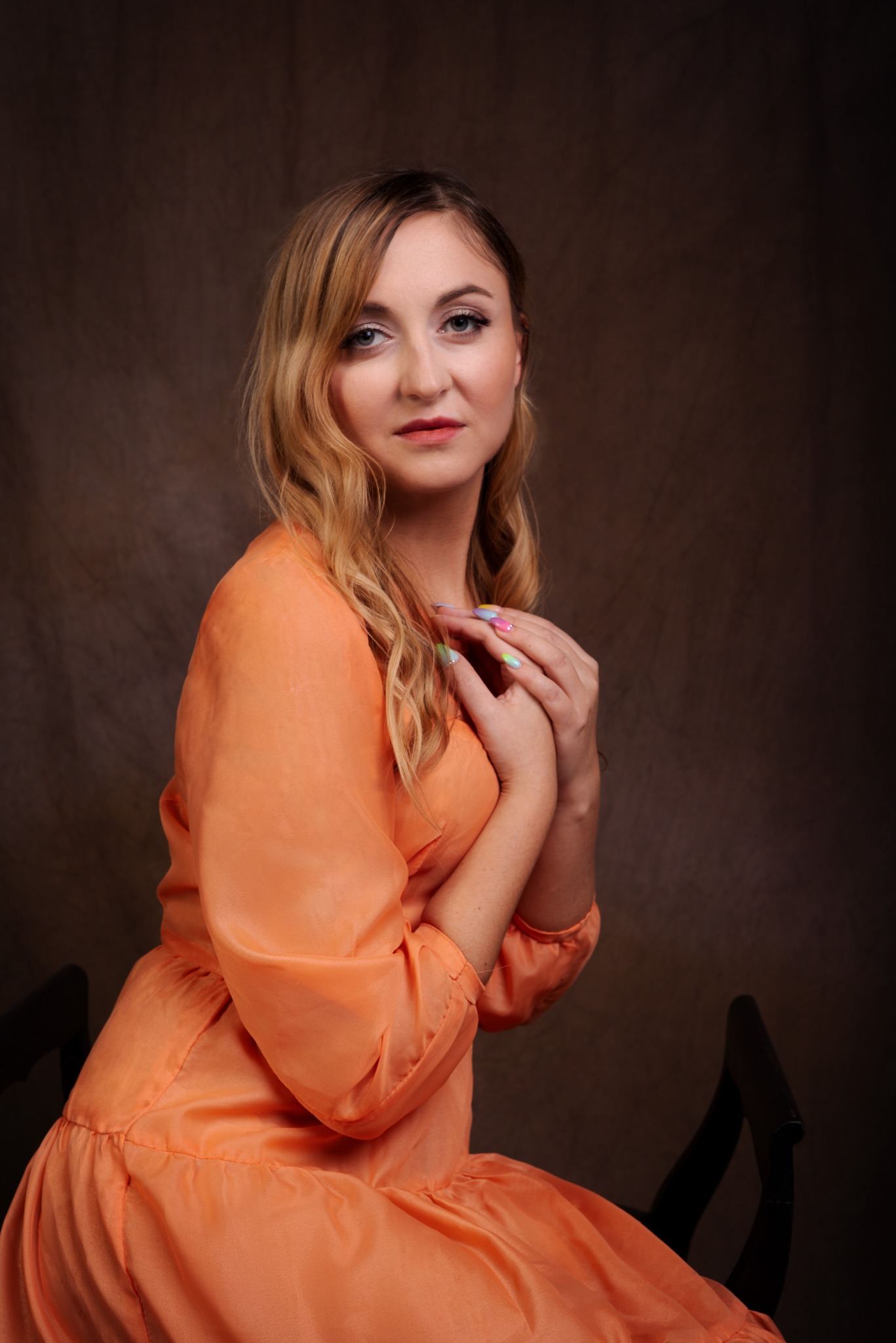 Portret kobiety w pomarańczowej sukience, z dłońmi złożonymi pod brodą, na ciemnobrązowym tle. Widoczne pastelowe zdobienia paznokci.