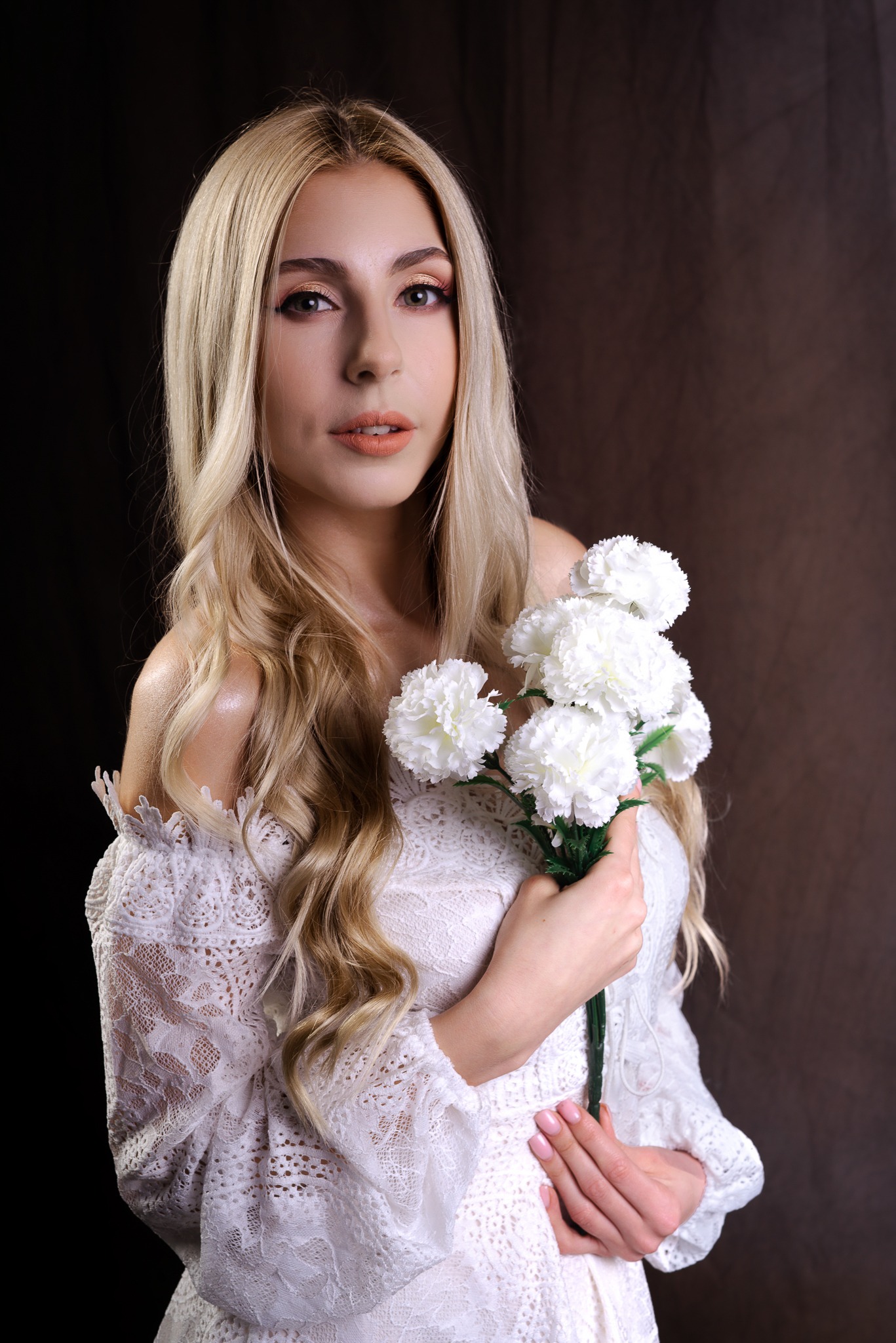 Blondwłosa kobieta w białej koronkowej sukience trzyma bukiet białych goździków na ciemnym tle.