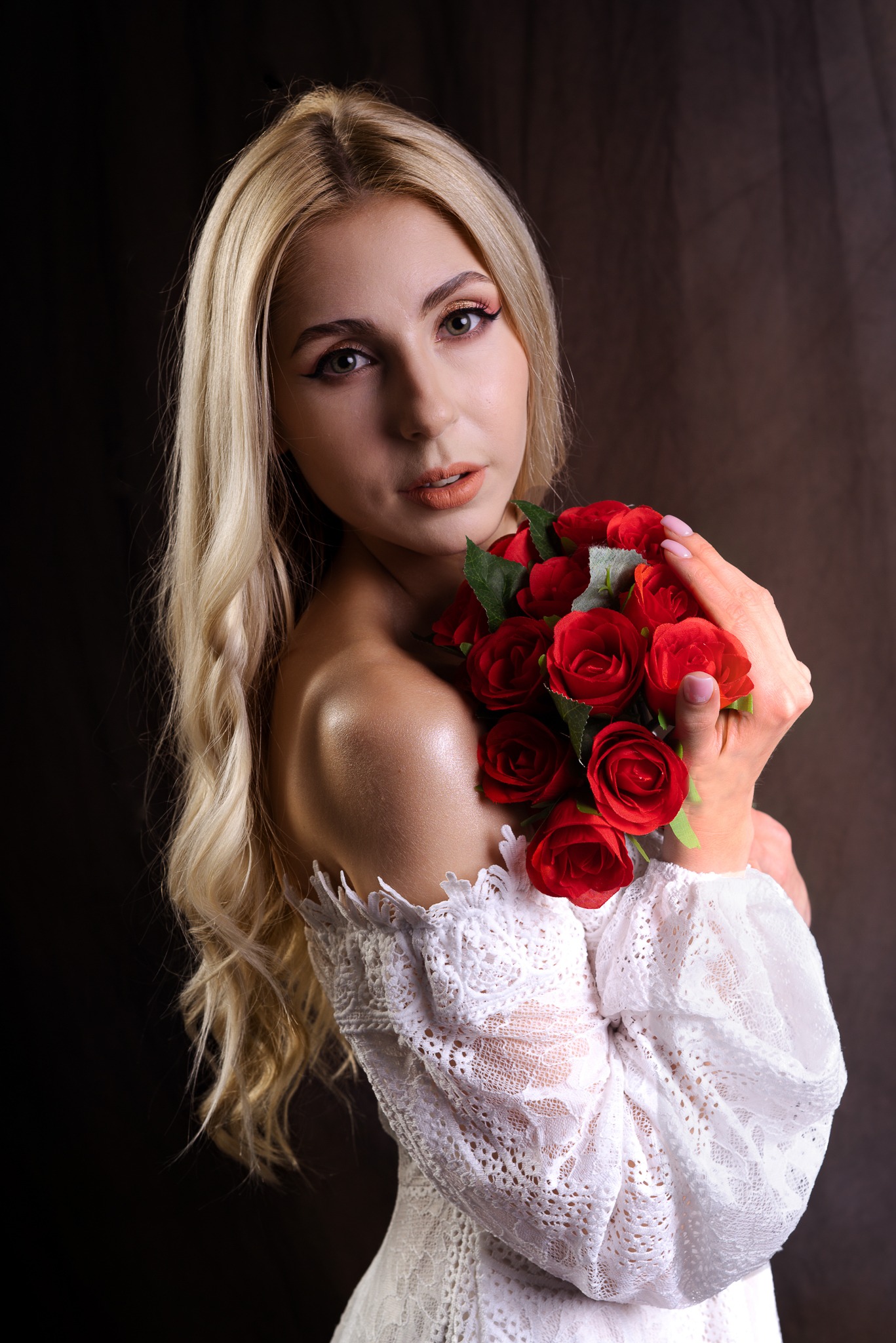 Portret kobiety z blond włosami, trzymającej bukiet czerwonych róż, ubrana w białą koronkową sukienkę na tle ciemnego, drapowanego materiału.