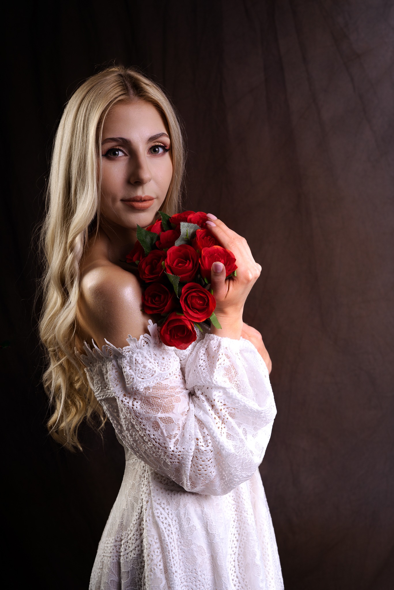 Portret kobiety z blond włosami w białej koronkowej sukience, trzymającej bukiet czerwonych róż na tle ciemnego, brązowego tła.
