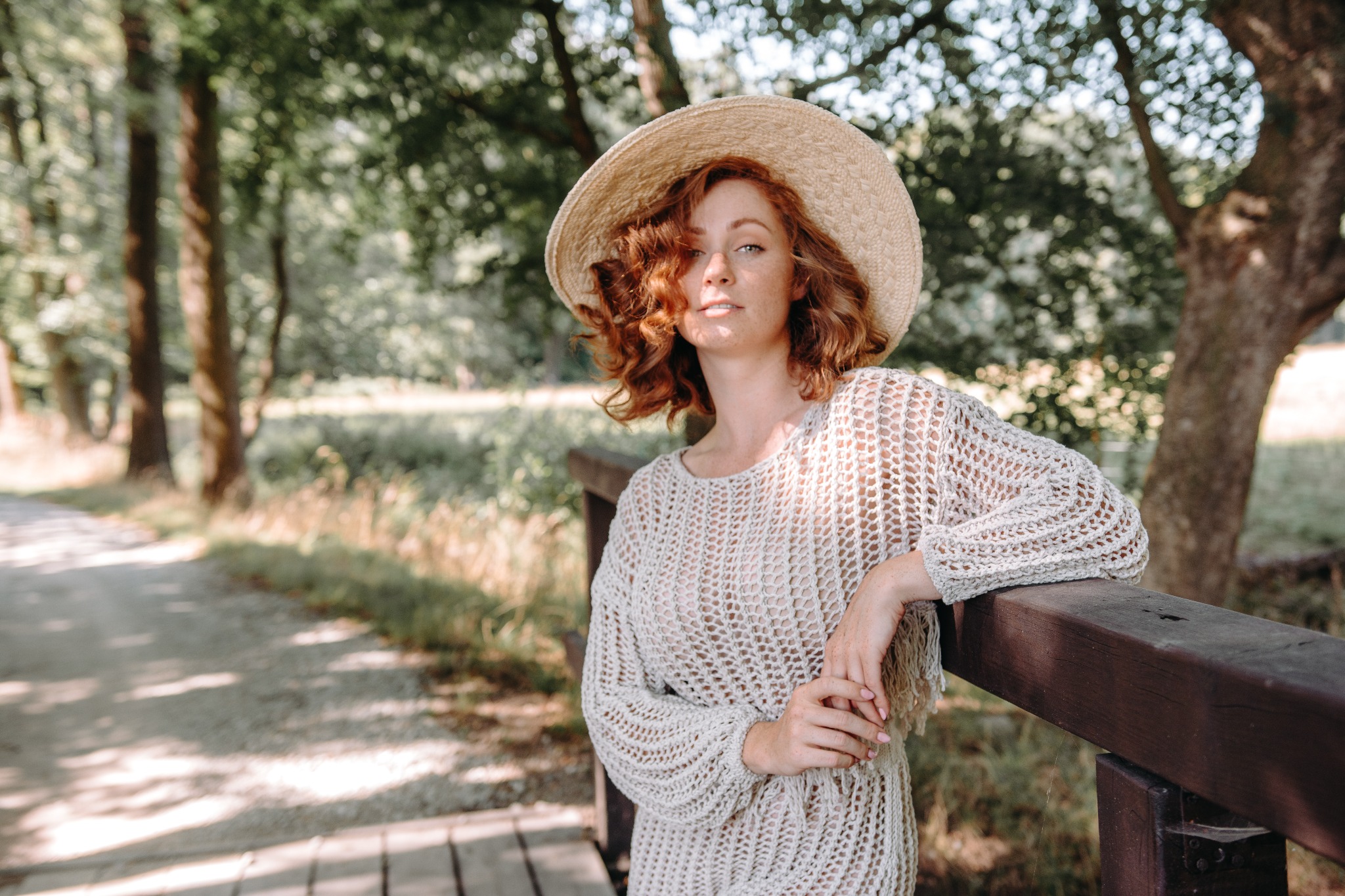 Kobieta z rudymi włosami i piegami w słomkowym kapeluszu i ażurowym swetrze opiera się o drewnianą barierkę na tle leśnej ścieżki.
