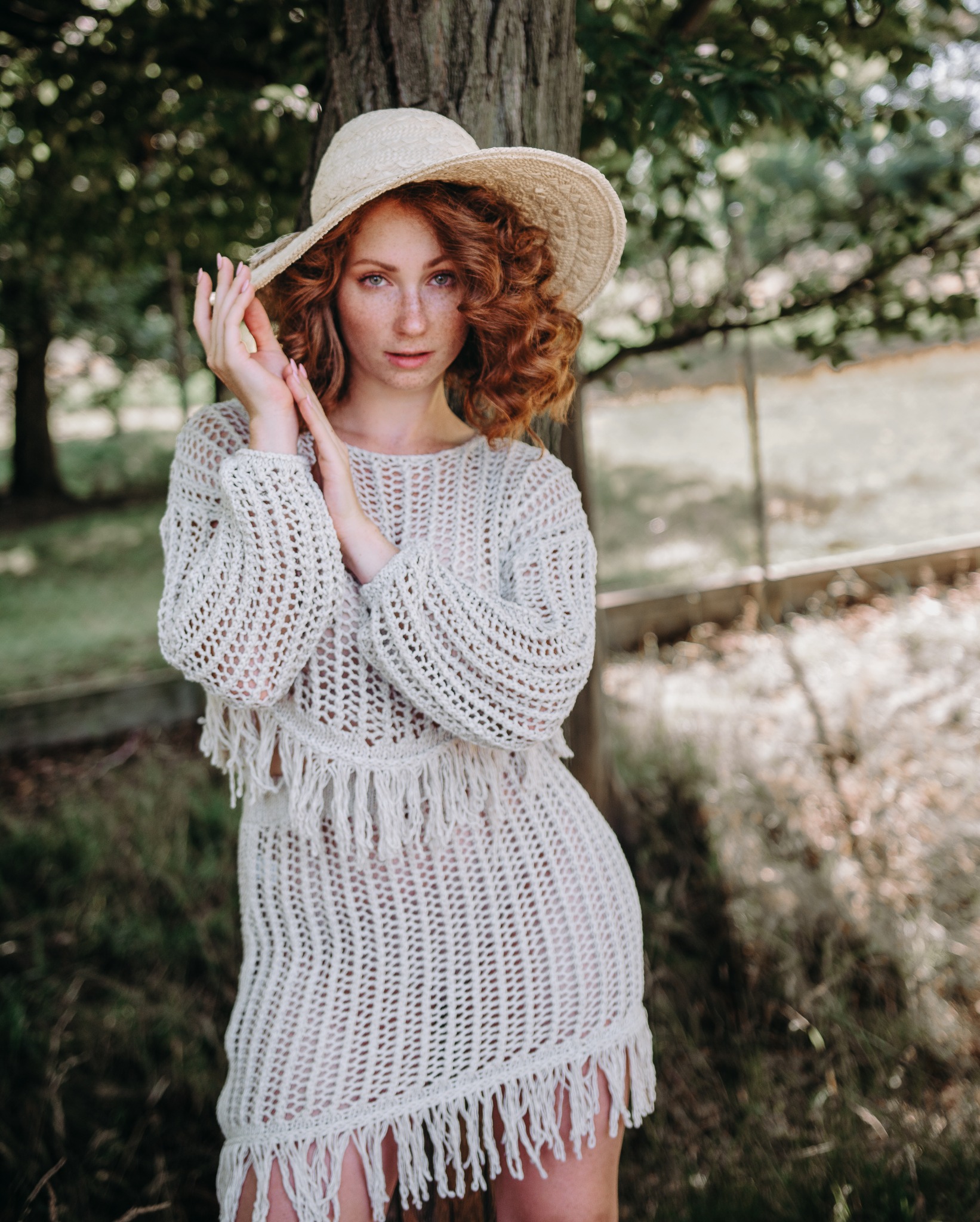 Portret rudej kobiety w ażurowej tunice i słomkowym kapeluszu, stojącej przy drzewie w plenerze, z dłońmi uniesionymi w kierunku twarzy.