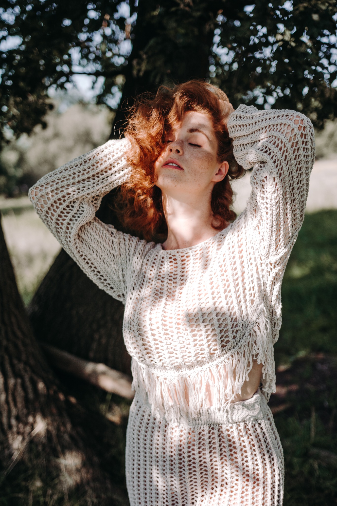 Rudowłosa kobieta w ażurowym stroju, stojąca pod drzewem z zamkniętymi oczami i rękami uniesionymi nad głową, w promieniach słońca.