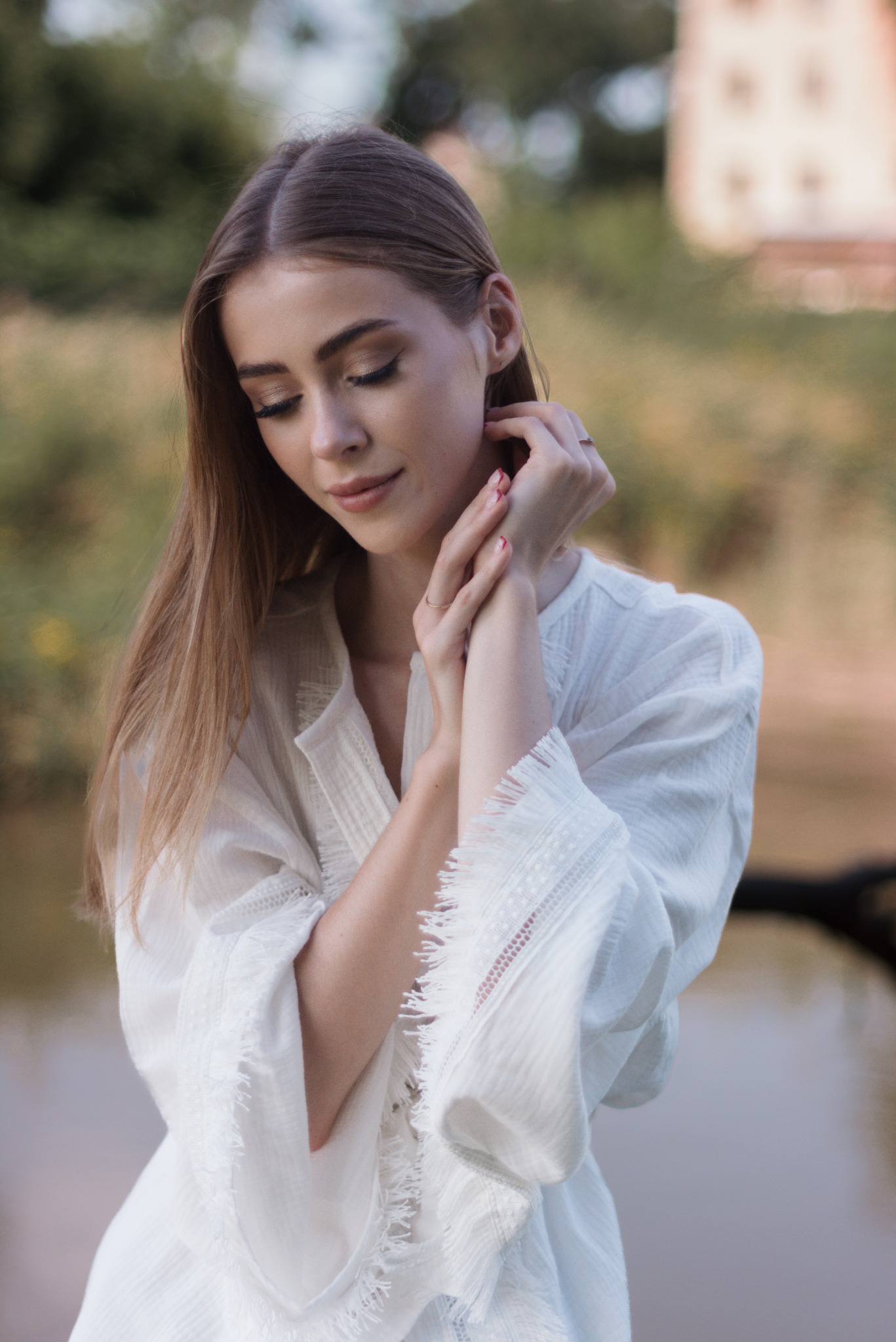 Portret młodej kobiety w białej, zwiewnej koszuli z koronkowymi detalami na tle rozmytej zieleni i wody, z delikatnym uśmiechem i splecionymi dłońmi, eksponujący subtelny makijaż i pierścionek.