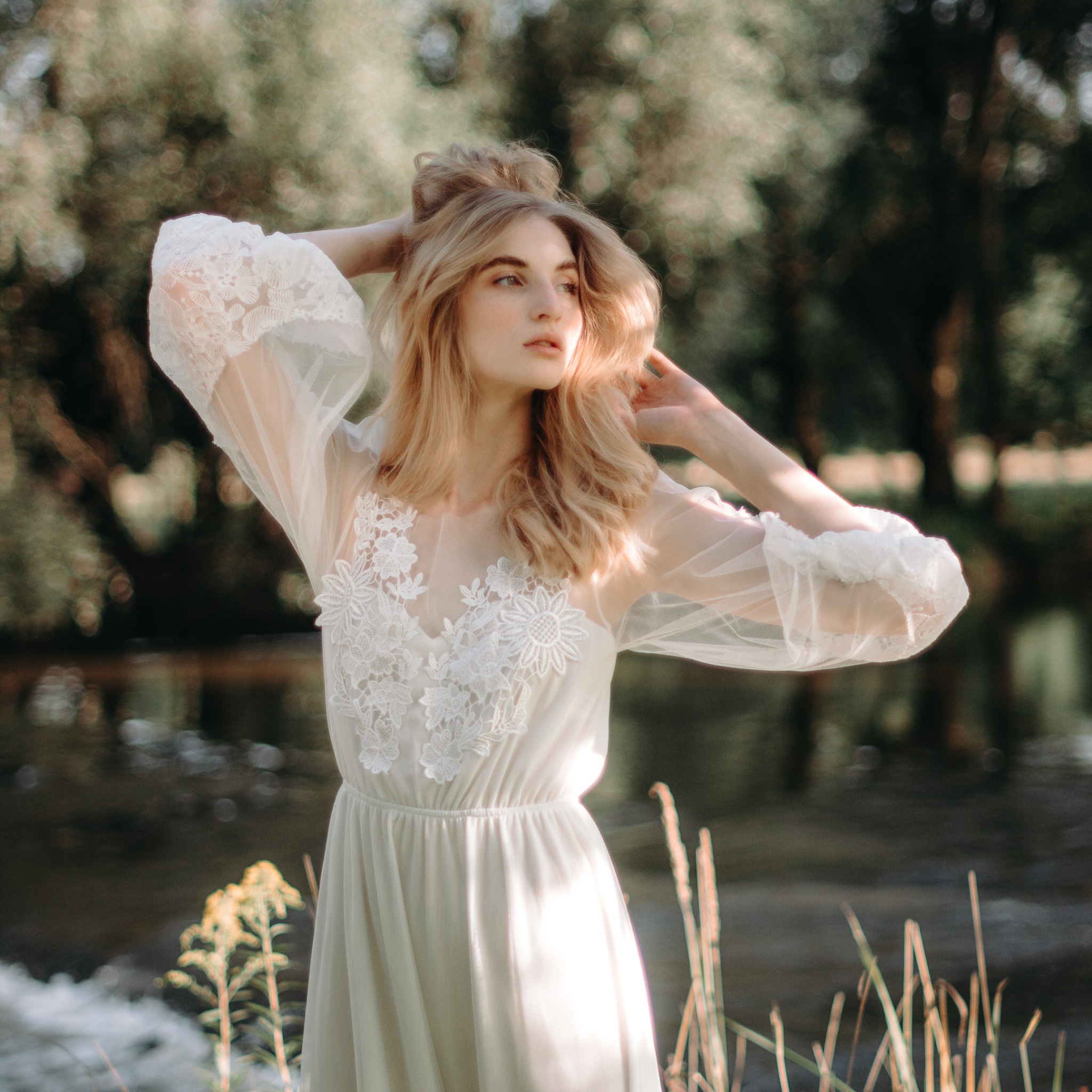 Kobieta w zwiewnej, koronkowej sukience pozuje nad rzeką, trzymając ręce za głową; miękkie, naturalne światło i rozmyte tło.