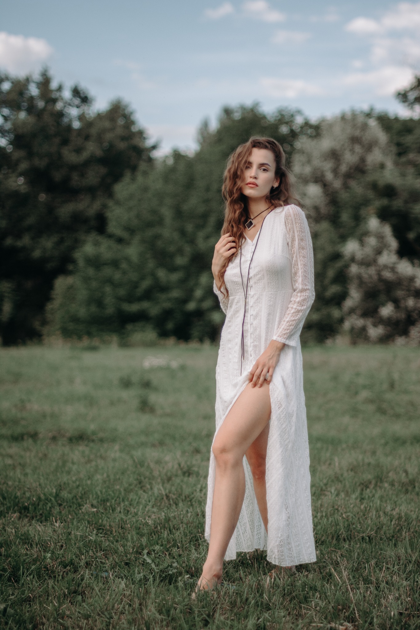 Kobieta w zwiewnej, białej sukience z koronkowymi rękawami, stojąca boso na trawie, z rozcięciem ukazującym nogę, pozująca na tle zieleni drzew i błękitnego nieba.