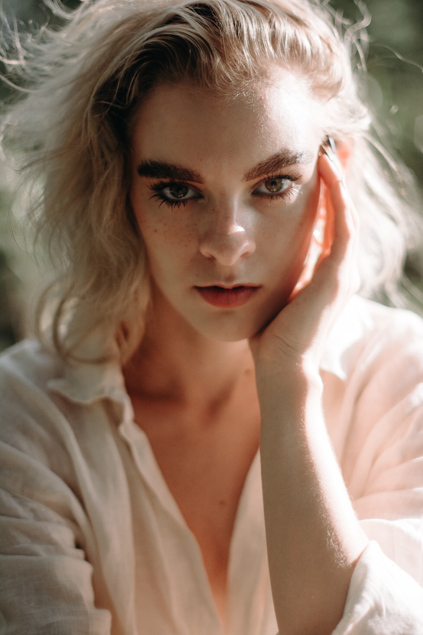 Portret kobiety z jasnymi włosami, delikatnym makijażem i piegami, w białej koszuli, pozująca w naturalnym świetle z dłonią przy twarzy.