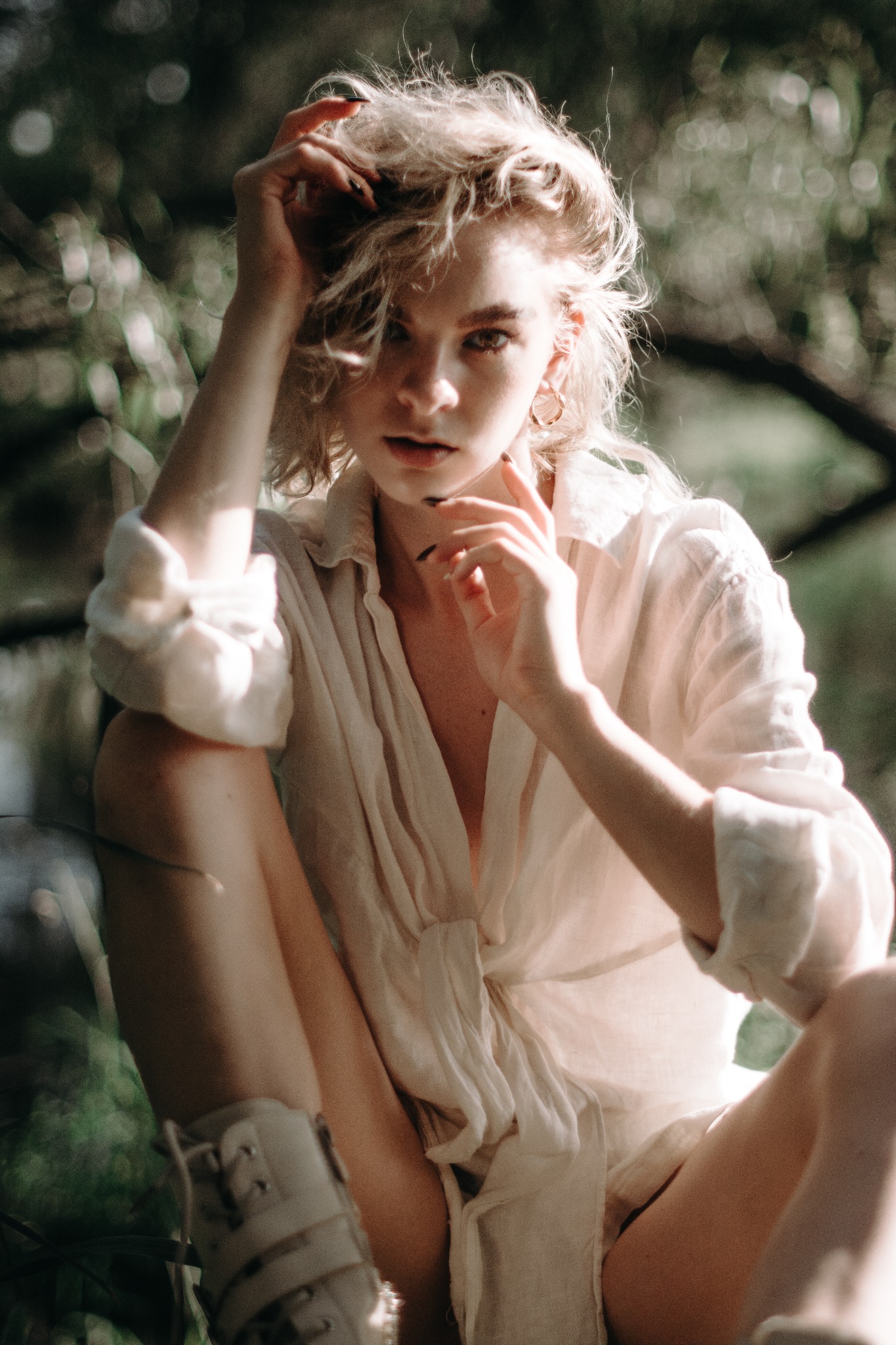 Portret młodej kobiety z blond włosami w rozpiętej, lnianej koszuli, siedzącej w plenerze z rękami uniesionymi do włosów, z delikatnym makijażem i złotymi kolczykami.