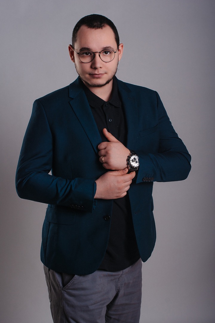 Mężczyzna w okularach i ciemnozielonej marynarce pozuje na szarym tle, poprawiając marynarkę i eksponując duży zegarek na nadgarstku.