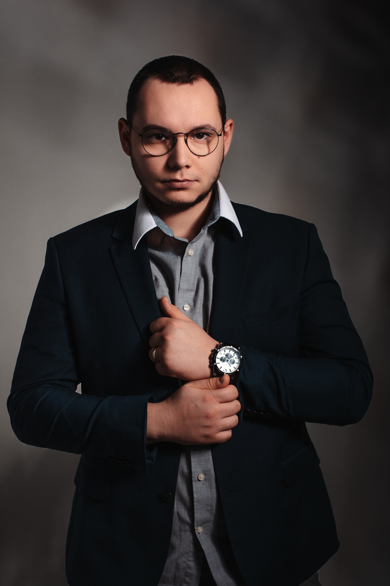 Portret mężczyzny w okularach, marynarce i koszuli, z zegarkiem na ręku, pozującego na szarym tle.