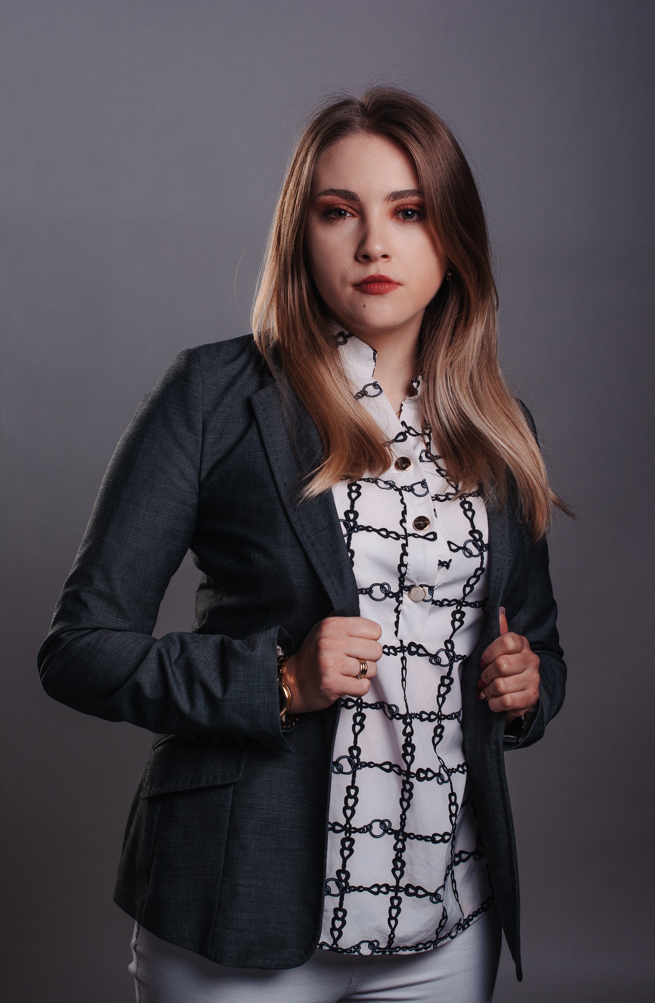 Portret kobiety w szarym żakiecie i bluzce w łańcuchy, złota bransoletka i pierścionek, neutralne tło, profesjonalny makijaż.