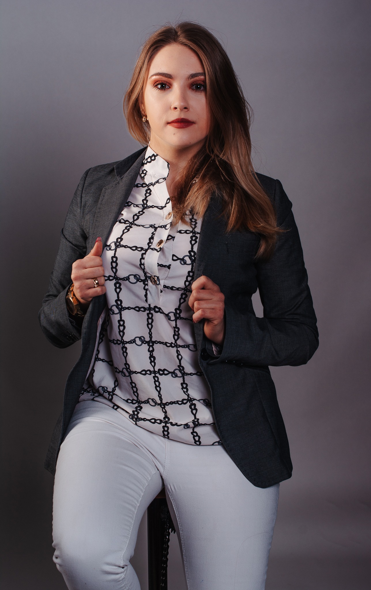 Portret kobiety w szarym żakiecie i białej bluzce w czarne łańcuchy, siedzącej na stołku na szarym tle, z delikatnym makijażem i złotym zegarkiem.