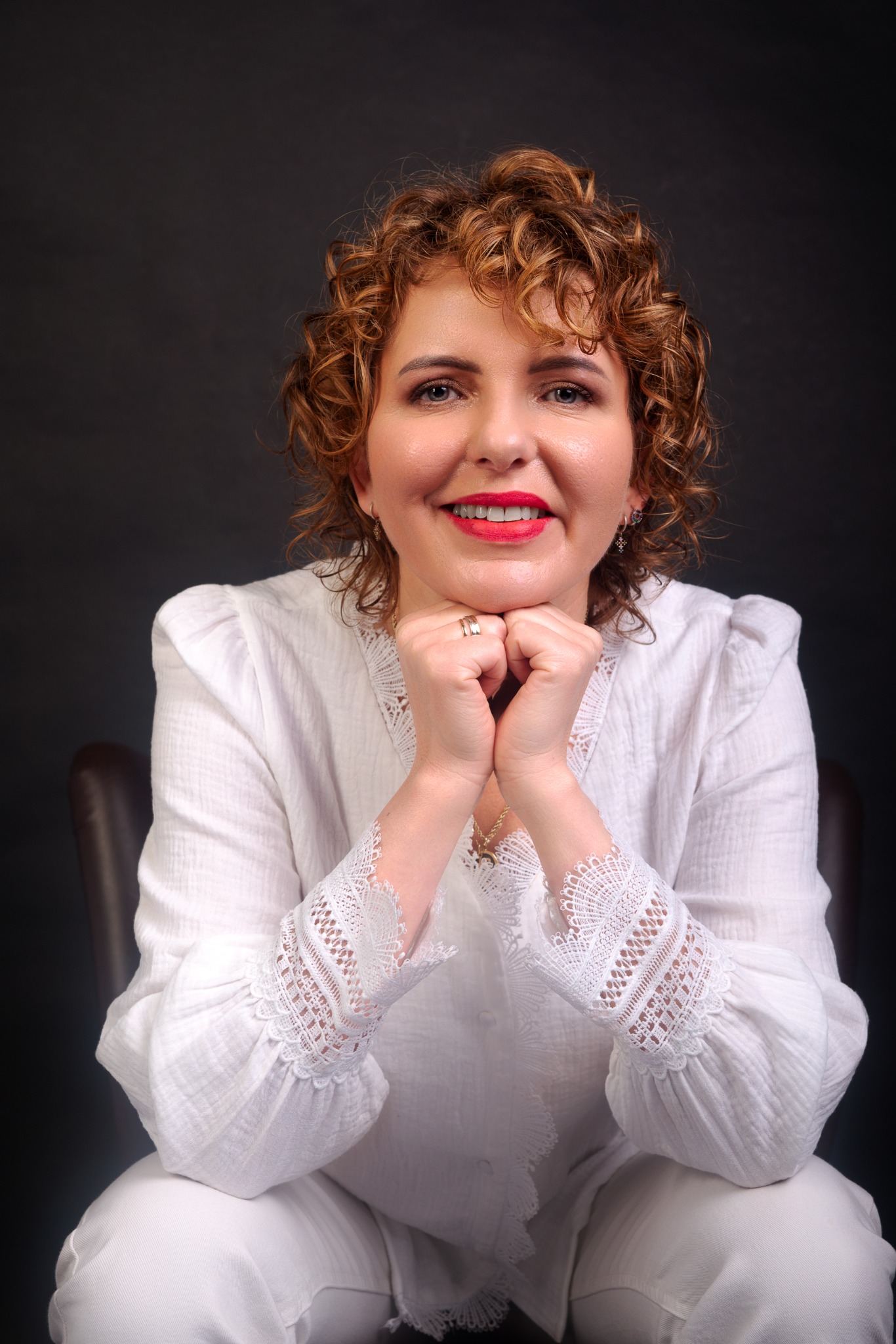 Portret uśmiechniętej kobiety z kręconymi rudymi włosami, opierającej podbródek na dłoniach, ubrana w białą bluzkę z koronkowymi rękawami, na ciemnoszarym tle.