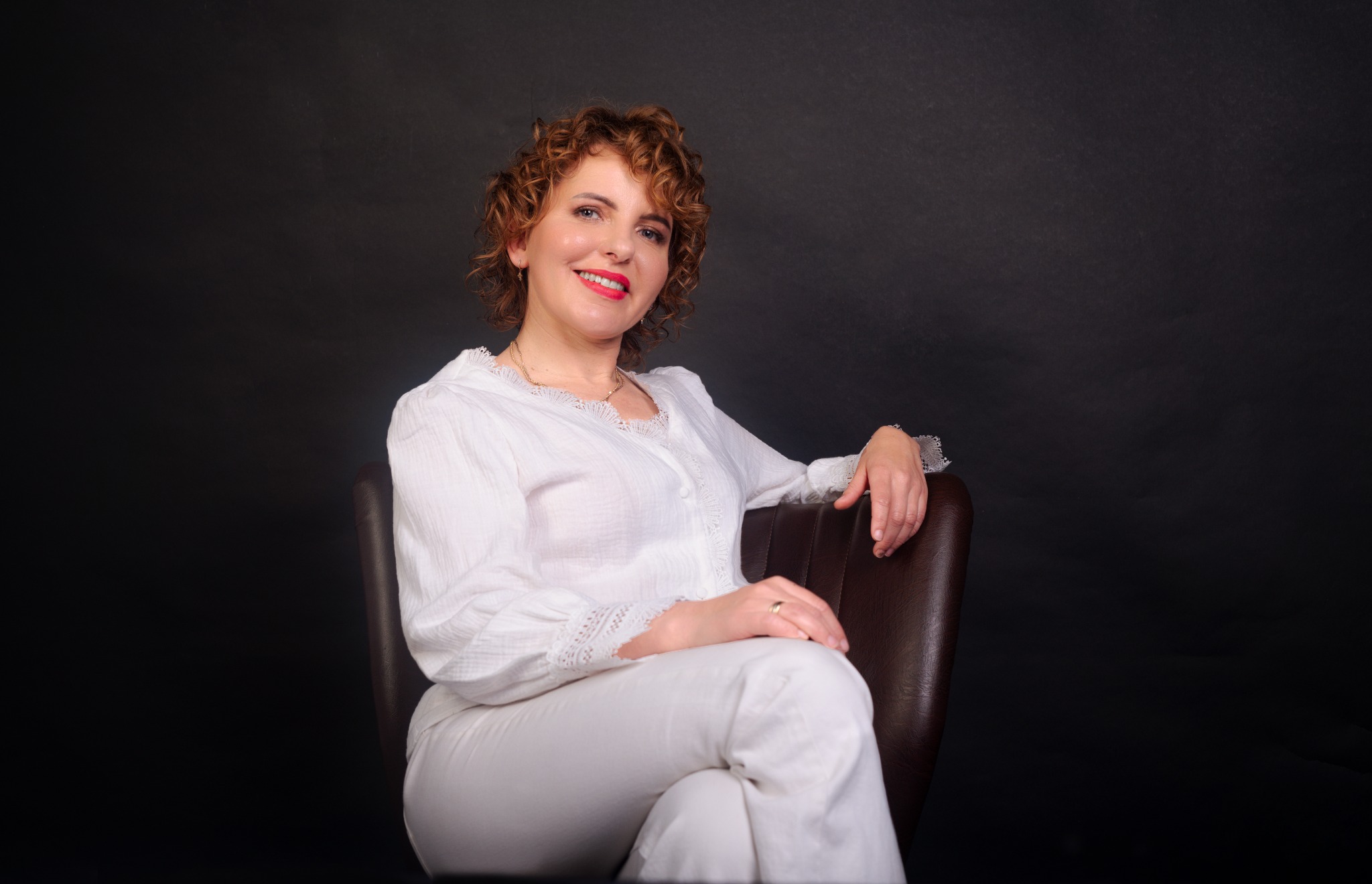Portret kobiety z kręconymi włosami w białym stroju, siedzącej w skórzanym fotelu na ciemnym tle. Uśmiech z czerwoną szminką, delikatna biżuteria.