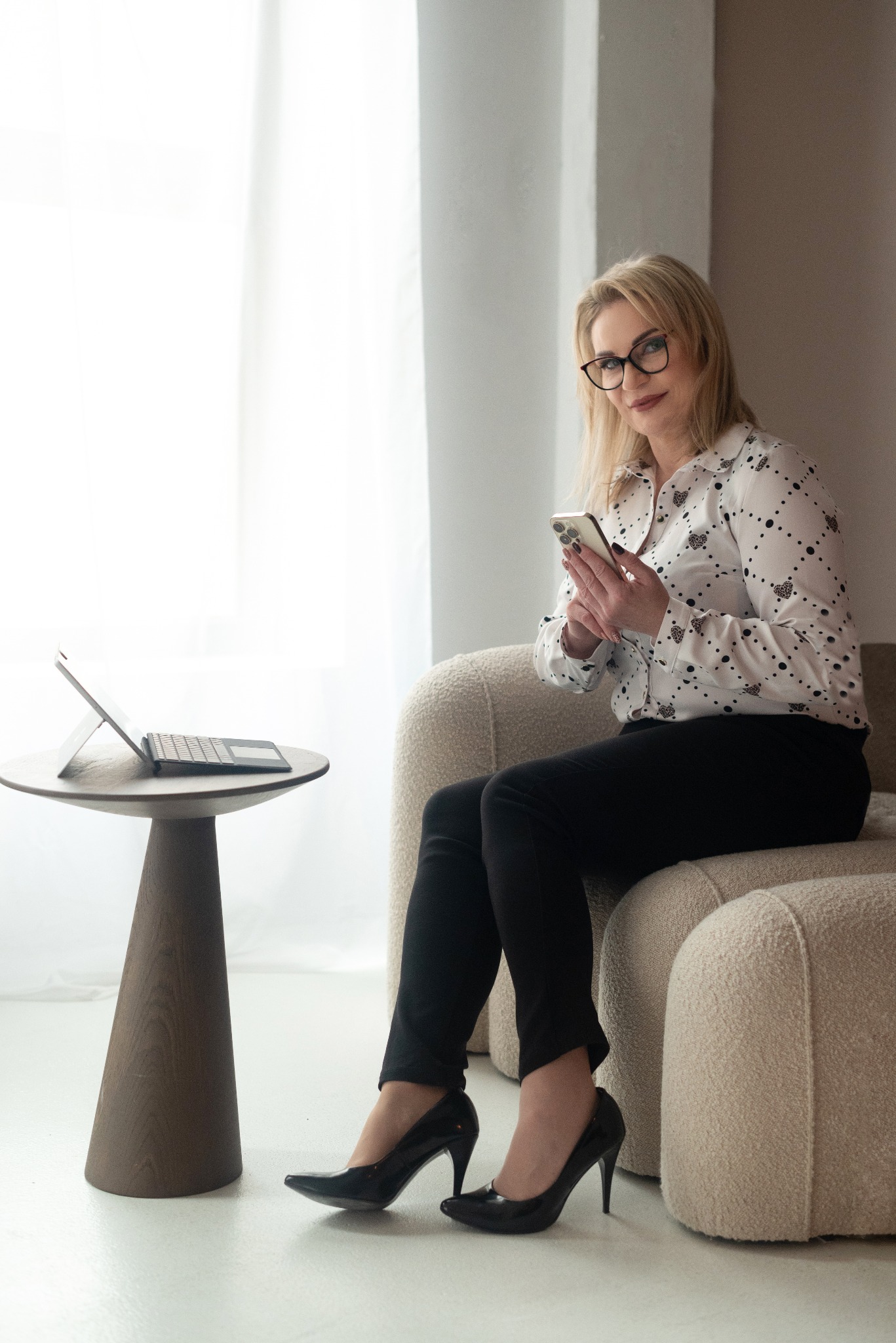 Kobieta w okularach siedzi na beżowym fotelu, trzymając smartfon, obok stolik z laptopem, jasne wnętrze.