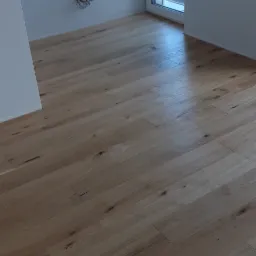 Jasne, lakierowane deski podłogowe z widocznymi sękami, ułożone w nowym budynku z białymi ścianami i oknem z widokiem na ulicę.