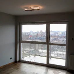 Pusty pokój po remoncie z drewnianą podłogą, grzejnikiem, dużym oknem balkonowym z widokiem na miasto i nowoczesnym oświetleniem sufitowym.