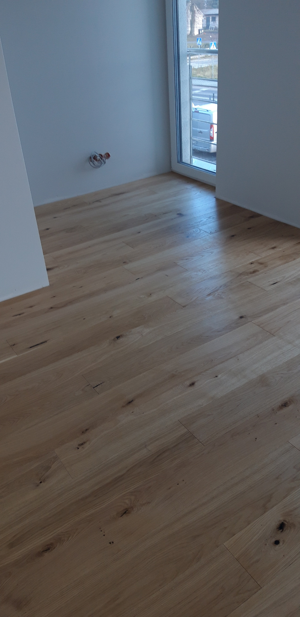Jasne, lakierowane deski podłogowe z widocznymi sękami, ułożone w nowym budynku z białymi ścianami i oknem z widokiem na ulicę.