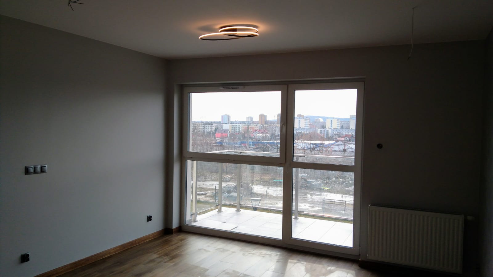Pusty pokój po remoncie z drewnianą podłogą, grzejnikiem, dużym oknem balkonowym z widokiem na miasto i nowoczesnym oświetleniem sufitowym.