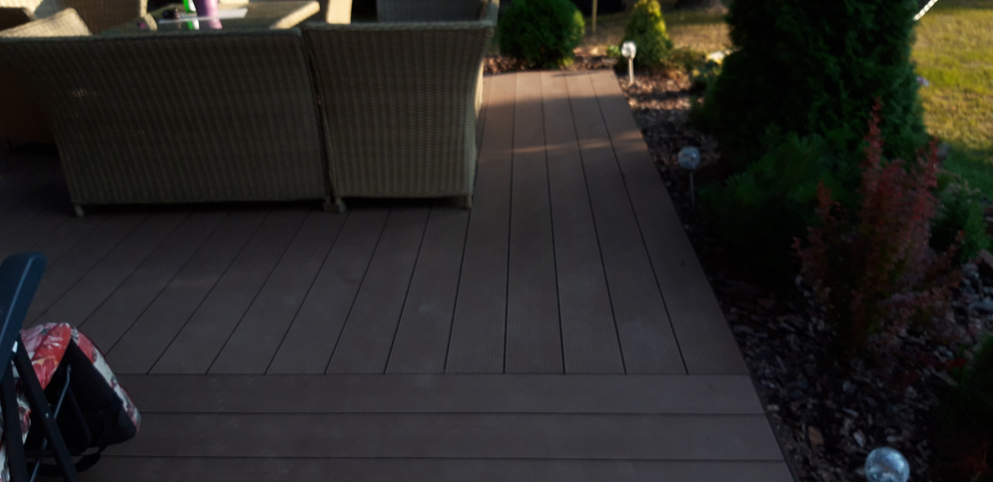 Kompozytowy taras z meblami ogrodowymi i oświetleniem wkomponowany w zieleń ogrodu.