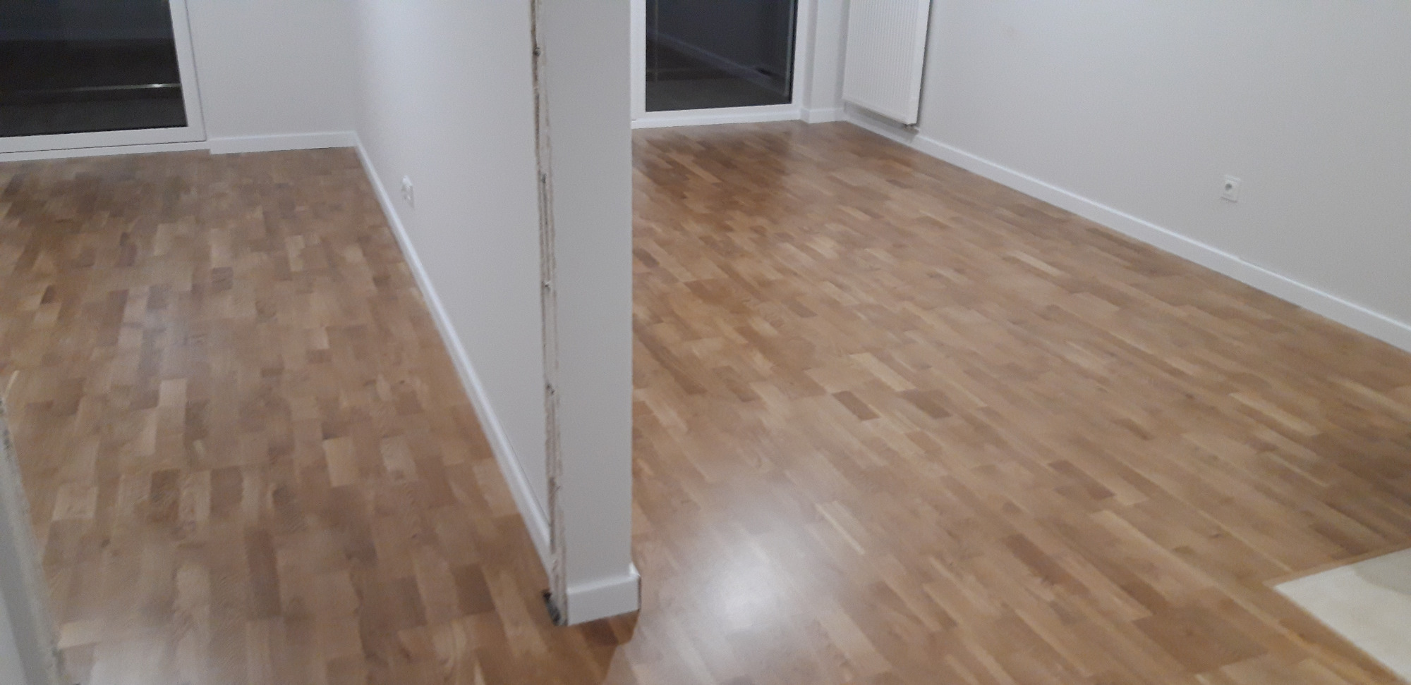 Jasnobrązowa podłoga z parkietu ułożona w jodełkę w pustym, jasnym pokoju z białymi ścianami i listwami przypodłogowymi.