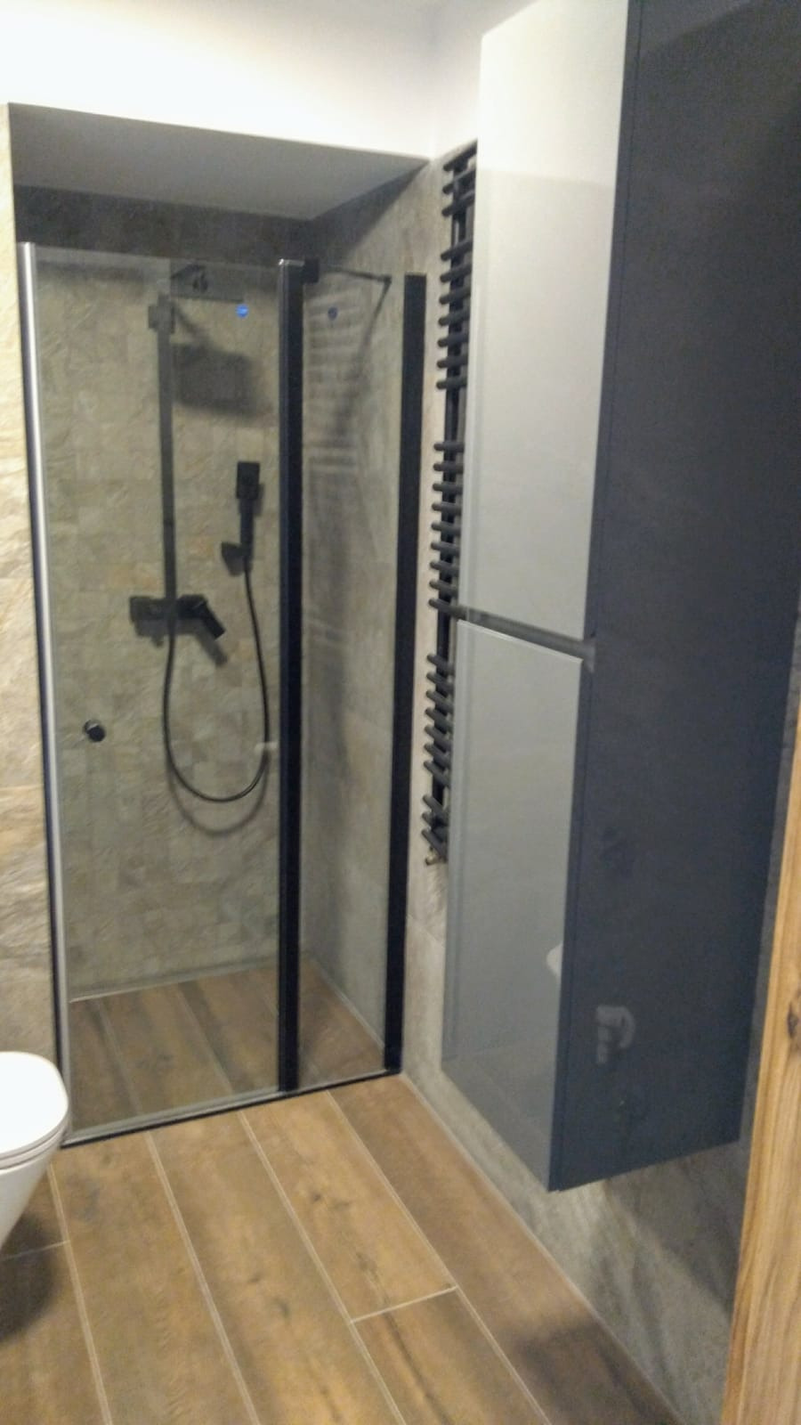 Nowoczesna łazienka z kabiną prysznicową z czarną armaturą, grzejnikiem drabinkowym i szafką w odcieniach szarości, widok na drewnopodobną podłogę.